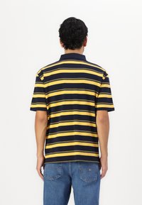 Polo shirt med marineblå og gule vandrette striber, korte ærmer og krave. Bæres med blå jeans, vist fra ryggen.