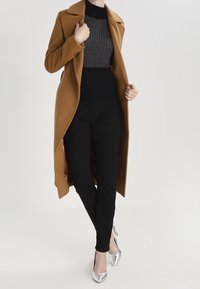Un manteau en camelle avec une finition texturée, un design long et des revers à cran, associé à un pull noir à pois blancs et un pantalon noir. Des talons argentés viennent accentuer la tenue.