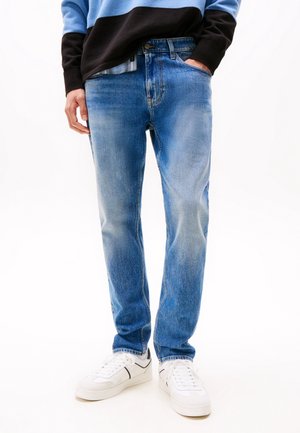 Personne portant un jean bleu délavé, un pull noir et bleu, des baskets blanches, avec une main dans la poche, sur un fond clair uni.
