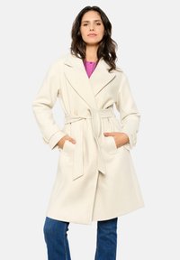 Cream-colored belted coat with a collar, two front pockets, and a textured fabric. Worn over a pink top and paired with blue jeans.