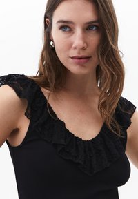 Musta ribattu tank top, jossa on pitsinen röyhelökaulus ja -olkaimet. Pehmeä tekstuuri ja tiukka siluetti. Näkyvät hopeiset korvakorut.