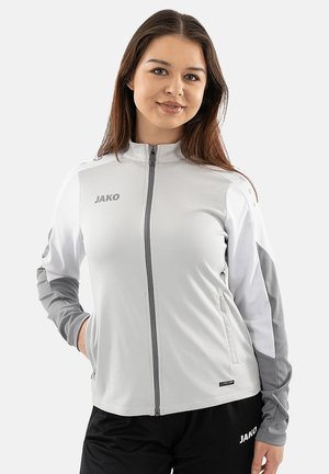 Donna che indossa una giacca sportiva con zip grigio chiaro e bianca con logo e pantaloni sportivi neri con logo, in piedi con una mano in tasca.