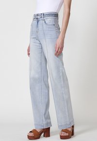 Jeans en denim bleu clair taille haute avec un détail tressé au niveau de la ceinture, coupe large et coutures visibles. Portés avec des sandales à plateforme marron.