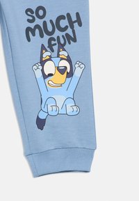Lyseblå joggebukser med ribbede mansjetter har et tegneseriehundmotiv og teksten "SÅ MYE MORO" i fet, svart skrift.