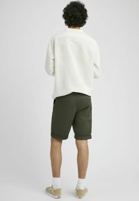 Chemise en lin blanche à manches longues, shorts roulés vert olive et baskets beiges. Détails visibles au dos comprenant des coutures et un design décontracté.