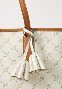 Sac crème avec un motif texturé, doté d'une bandoulière marron et de franges décoratives. Le design met en avant l'élégance et la sophistication.