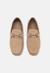 Mocasines de ante color beige con herrajes plateados decorativos, detalles de costura y suela plana. Diseñados con una forma clásica de mocasín.