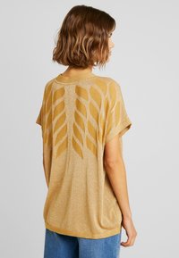 Mujer con cabello castaño ondulado que lleva un suéter corto de manga corta, suelto y dorado, con un patrón de hojas en la espalda, combinado con jeans azules.