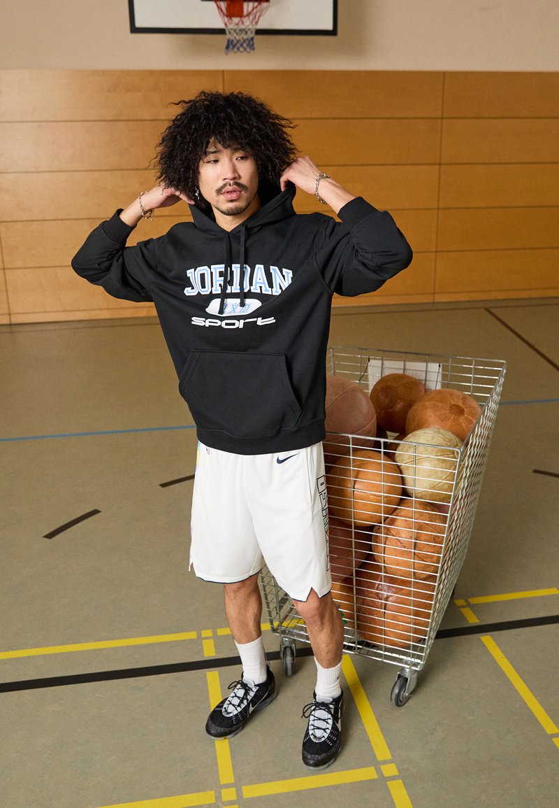 Sudadera negra con "JORDAN" en azul claro, pantalones cortos blancos, zapatillas negras, de pie junto a un carro de alambre lleno de balones de baloncesto marrones.