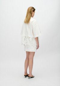 Blouse blanche à manches amples et finition en dentelle, associée à une mini-jupe en dentelle blanche. Le mannequin porte des talons noirs, affichant une esthétique minimaliste.