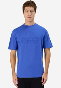 Camiseta de algodón azul con cuello redondo, mangas cortas y un logotipo texturizado de Kappa en el frente. Paneles laterales en amarillo contrastante.