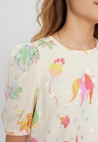 Kraschblus i krämfärg med flerfärgade mönster och metalliska prickar. Har puffärmar och rund halsringning.