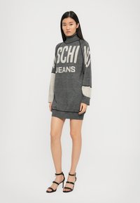 Pelēks adīts kleita ar "Moschino Jeans" logotipu, augsts kapuci, garām piedurknēm un kontrastējošām gaiši pelēkām akcentiem pie piedurknēm.