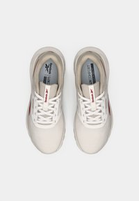 Zapatillas deportivas blancas con un upper texturizado, acentos en gris, logotipos rojos y una suela acolchada. Los cordones son blancos, con un diseño simil malla.