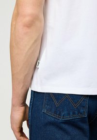 Vit bomullst-shirt med en etikett vid fållen, kombinerad med mörkblå denimjeans som har tydliga gula sömmar på bakfickan.