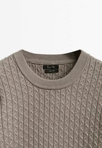 Maglione grigio lavorato a maglia con un motivo a treccia testurizzato, scollatura a coste e un piccolo etichetta interna che mostra il nome del marchio.