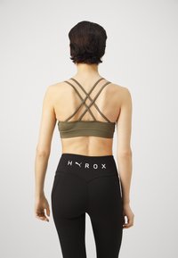 Sujetador deportivo verde oliva con tirantes entrecruzados, combinado con leggings negros de cintura alta que presentan el logo blanco "HYROX" en la cinturilla.