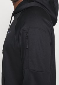 Sweatshirt à capuche noir avec une poche zippée sur le haut du bras, en tissu texturé. Logo Nike en bleu foncé sur la poitrine.