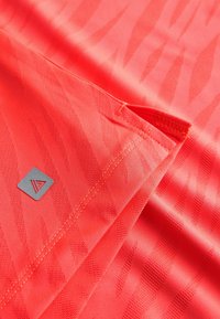Tissu corail vif avec un motif subtil de rayures de tigre, plié avec des coutures visibles et un petit patch logo triangulaire.