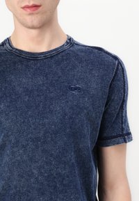 Tricou din bumbac de culoare albastru închis cu guler rotund, mâneci scurte și un logo discret pe piept. Prezintă o textură uzată și cusături în contrast.
