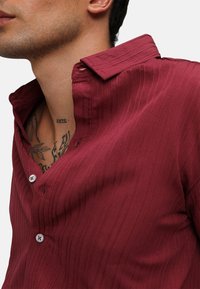 Camicia bordeaux in tessuto testurizzato, con patta dei bottoni e un sottile design a coste. Il colletto è leggermente aperto.