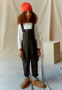 Sorte denim overalls med justerbare stropper, beige langærmet skjorte, orange ribbet hue, brune bløde støvler og grå sokker.
