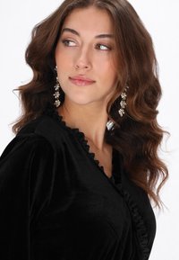 Fekete velúr blúz fodros nyakkivágás részlettel, lógó fülbevalóval viselve. A modell hullámos barna hajjal és természetes sminkkel rendelkezik.