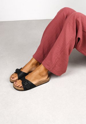 Schwarze Badesandalen mit Korksohle, die ein breites Band und eine verstellbare Schnalle haben. Getragen mit lockeren, rosa Hosen. Glatte Textur, schlichtes Design.