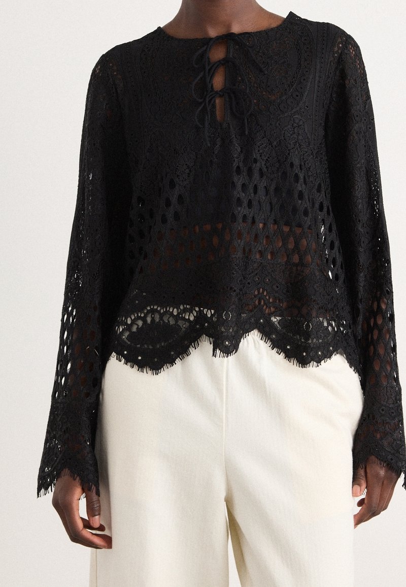 Blouse noire en dentelle à manches longues avec fermeture par lien et ourlet festonné, portée avec un pantalon blanc taille haute sur une personne à la peau foncée.