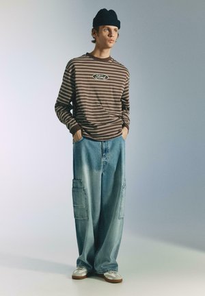 Braun-beiges, gestreiftes Langarmshirt mit "Ford"-Logo, kombiniert mit lockeren, hellblauen Denim-Cargohosen und weißen Sneakers. Dazu wird eine schwarze Mütze getragen.