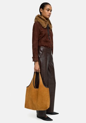 Brązowa kurtka zamszowa z kołnierzem z sztucznego futra, brązowe skórzane spodnie o szerokich nogawkach oraz beżowa torebka zamszowa z czarnymi wykończeniami, noszone przez stojącą postać.