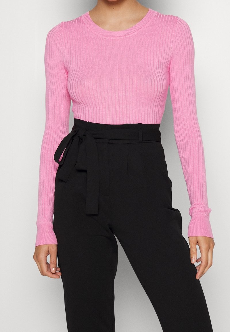 Femme portant un haut rose moulant à manches longues côtelées, rentré dans un pantalon noir taille haute avec une ceinture nouée à la taille.