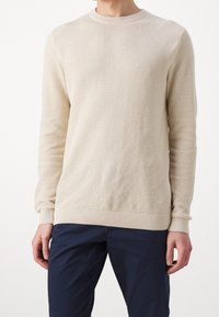 Beige stickad tröja med ett strukturerat mönster, ribbad krage och mudd, långa ärmar, i kombination med marinblå byxor.