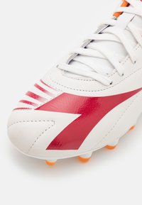 Futbalová obuv s bielym koženým exteriérom, červeným akcentom, prúžkovaným vzorom a oranžovými kolíkmi na päte. Viditeľné detaily šnurovania.