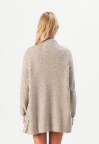 Blonde Frau von hinten zu sehen, die einen übergroßen beigen Strickpullover mit langen Ärmeln trägt, vor einem schlichten weißen Hintergrund.