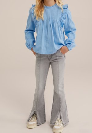 Niño vestido con una blusa de manga larga con volantes en azul claro, jeans grises de campana con aberturas delanteras y zapatillas blancas beige, con las manos en los bolsillos.