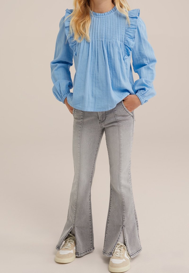 Enfant portant un blouse à volants bleu clair à manches longues, un jean grisé évasé avec des fentes à l'avant, et des baskets beige et blanches, les mains dans les poches.