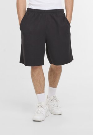 Mand iført sorte teksturerede shorts med hænderne i lommerne, hvide strømper og hvide sneakers, der står på en ensfarvet lys baggrund.