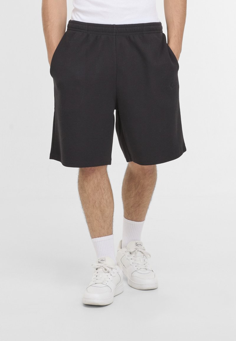 Homme portant un short noir texturé avec les mains dans les poches, des chaussettes blanches et des baskets blanches, debout sur un fond clair uni.