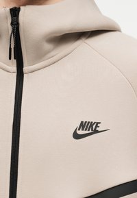 Close-up van een beige Nike zip-up hoodie met een zwarte rits en het Nike-logo op de borst.