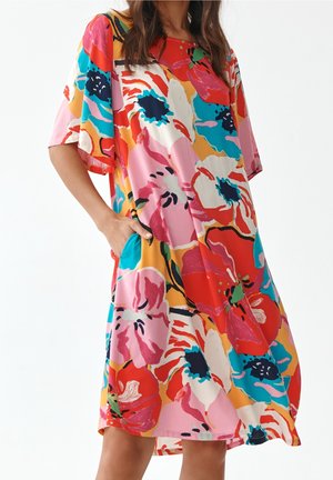 Kobieta w luźnej, sięgającej do kolan sukience z dużymi kolorowymi motywami kwiatowymi w kolorze czerwonym, różowym, niebieskim, białym i żółtym, z ręką w bocznej kieszeni.