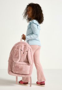 Lyserød plysbunny rygsæk med lange ører, frontlomme med lynlås og et smilende ansigt. Barnet har en lyseblå trøje og lyserøde leggings på.