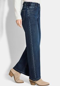 Femme portant un jean large bleu marine, un top à manches longues écru et des bottines en suède beige avec des talons en bois.