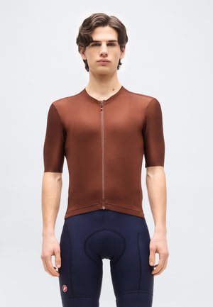 Νέος άνδρας με κυματιστά καστανά μαλλιά, φορώντας εφαρμοστό καφέ κοντομάνικο ποδηλατικό μπλουζάκι και ναυτικά μαξιλαρωμένα ποδηλατικά σορτς, όρθιος προς τα εμπρός.