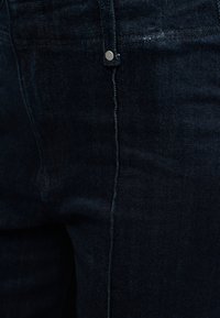 Κοντινό πλάνο σκούρου μπλε τζιν τζιν που δείχνει την μπροστινή τσέπη, τη ραφή και λεπτομέρειες με μεταλλικό κούμπωμα.