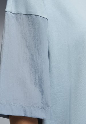 Haut bleu clair à manches courtes présentant deux textures de tissu différentes : une partie en maille lisse et une autre en tissu tissé froissé sur la manche.