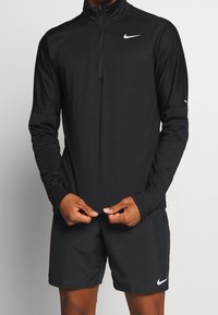 Maglietta nera a maniche lunghe Nike con una zip a quarto e una texture liscia, indossata con pantaloni corti neri abbinati con un piccolo logo sulla gamba.