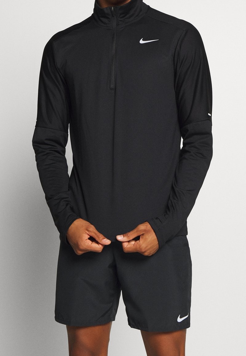 Maglietta nera a maniche lunghe Nike con una zip a quarto e una texture liscia, indossata con pantaloni corti neri abbinati con un piccolo logo sulla gamba.