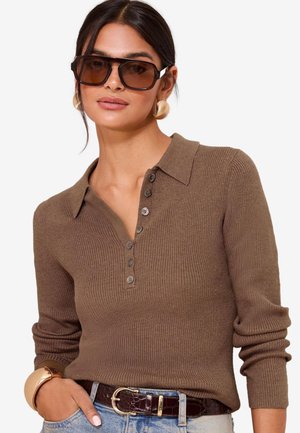 Donna con maglione polo marrone a coste a maniche lunghe, grandi occhiali da sole scuri, orecchini a cerchio dorati, bracciale rigido e jeans azzurri con cintura scura.