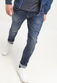 Jeans oscuros de corte ajustado con ligero desgaste, diseño de cinco bolsillos y puños enrollados, combinados con zapatillas blancas.
